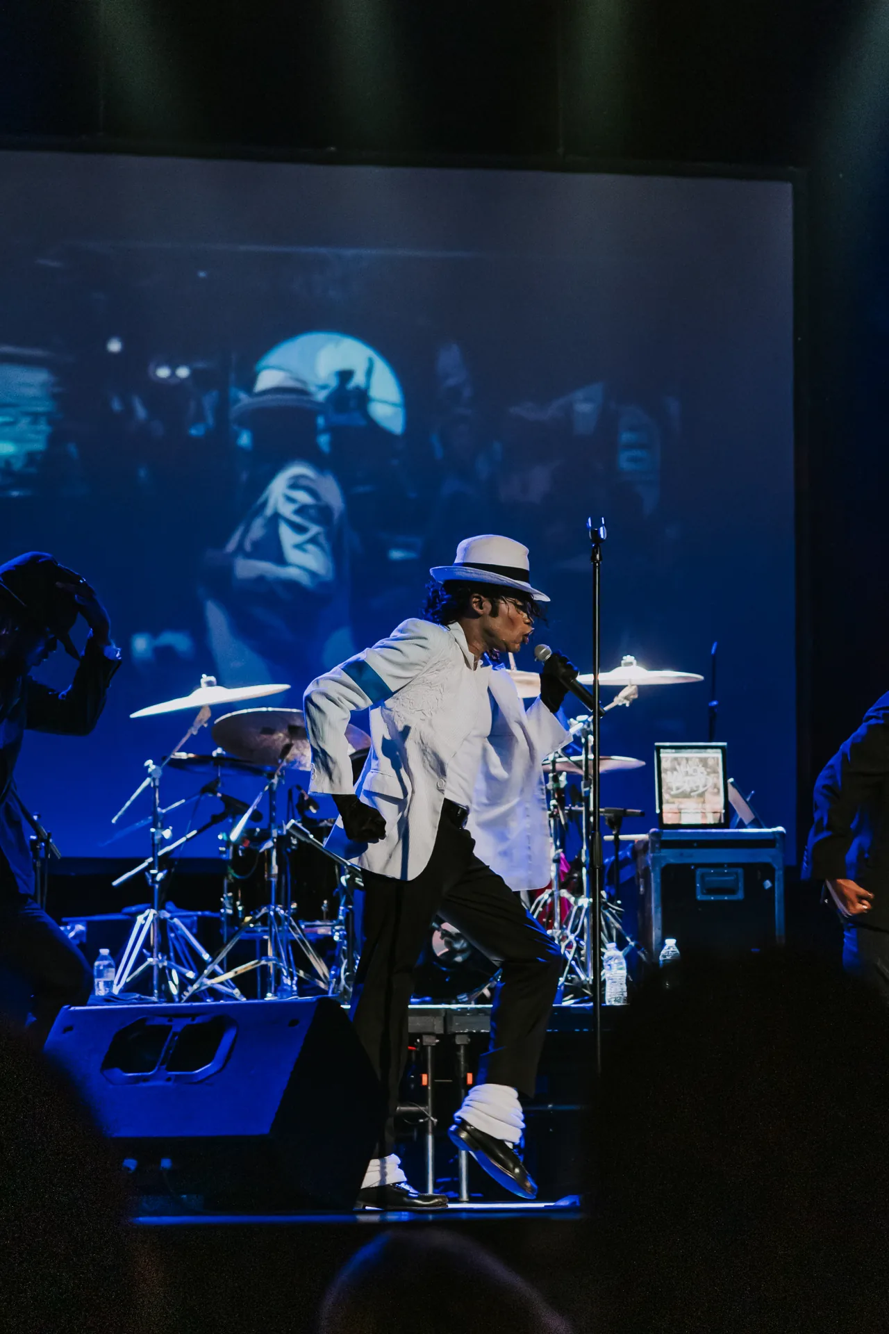 Who’s Bad vocalist in a white jacket sings in front of a drum kit under blue lighting