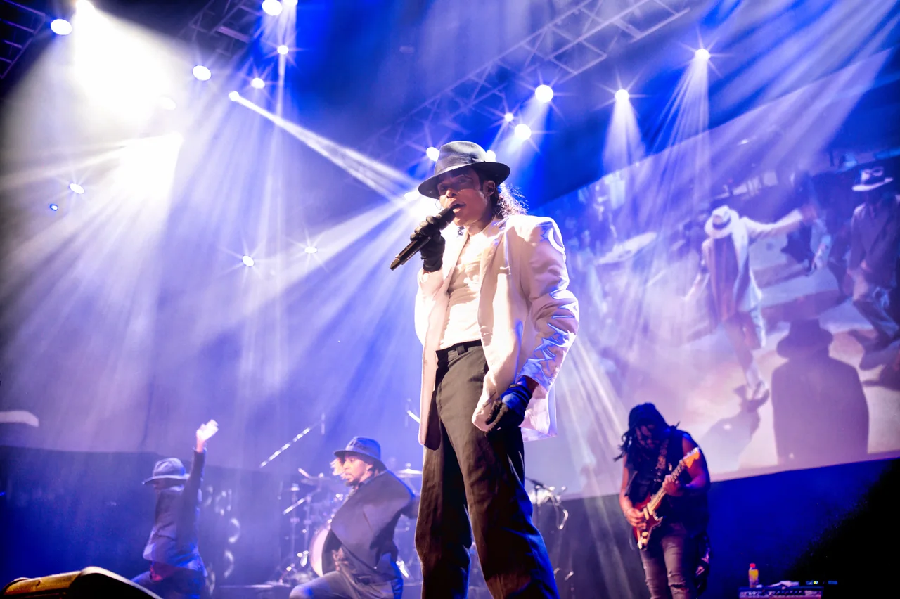 Who’s Bad performer in a white jacket and fedora sings with the band under blue and white stage lights